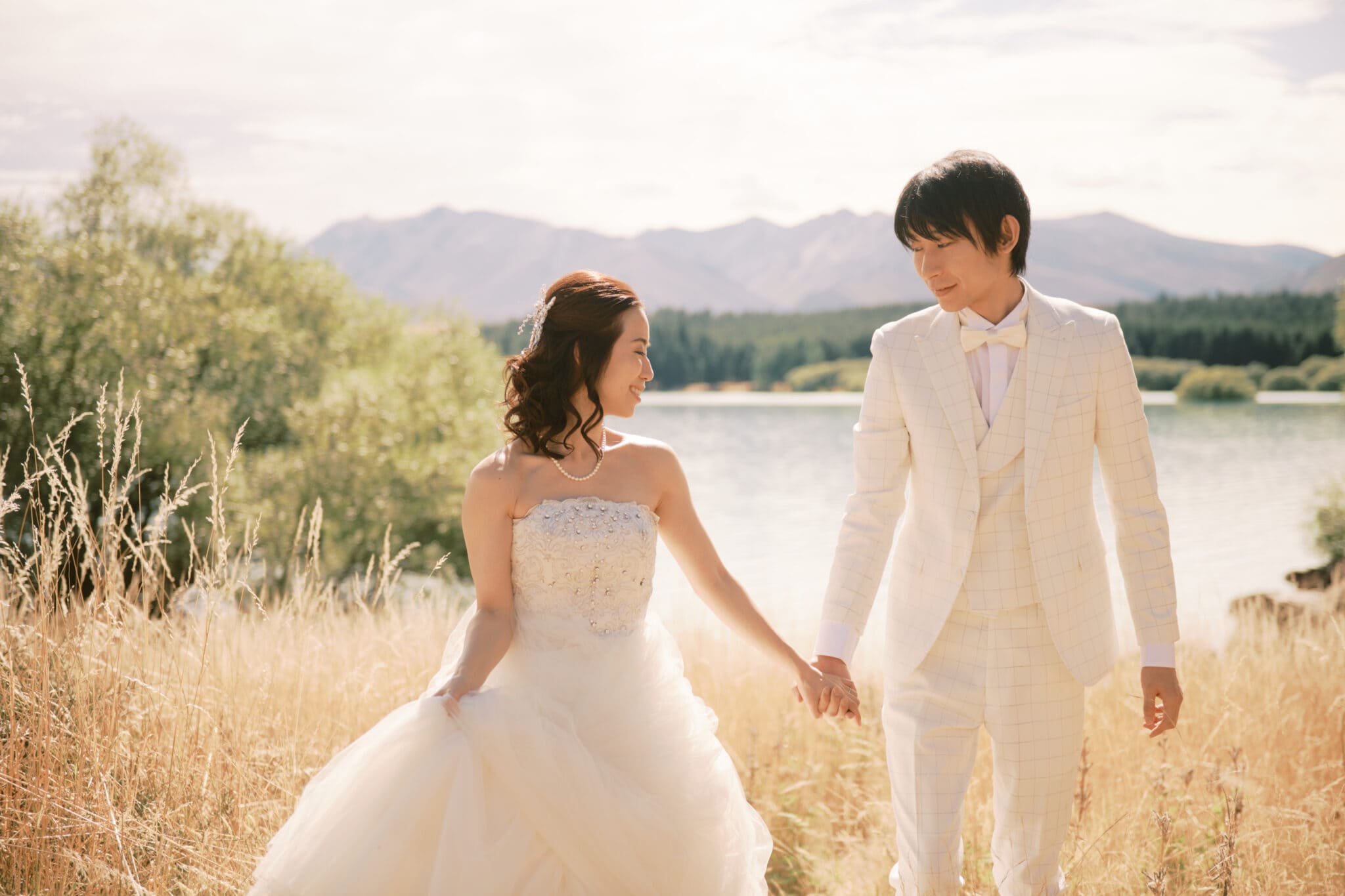 Queenstown Tekapo New Zealand Heli Wedding Elopement Pre-Wedding Shoot Photographer クイーンズタウン　テカポ　ニュージーランド　エロープメント 前撮り　フォトウェディング　結婚式 | A couple in wedding attire holding hands with a scenic lake and mountains in the background during their Tekapo pre-wedding shoot.